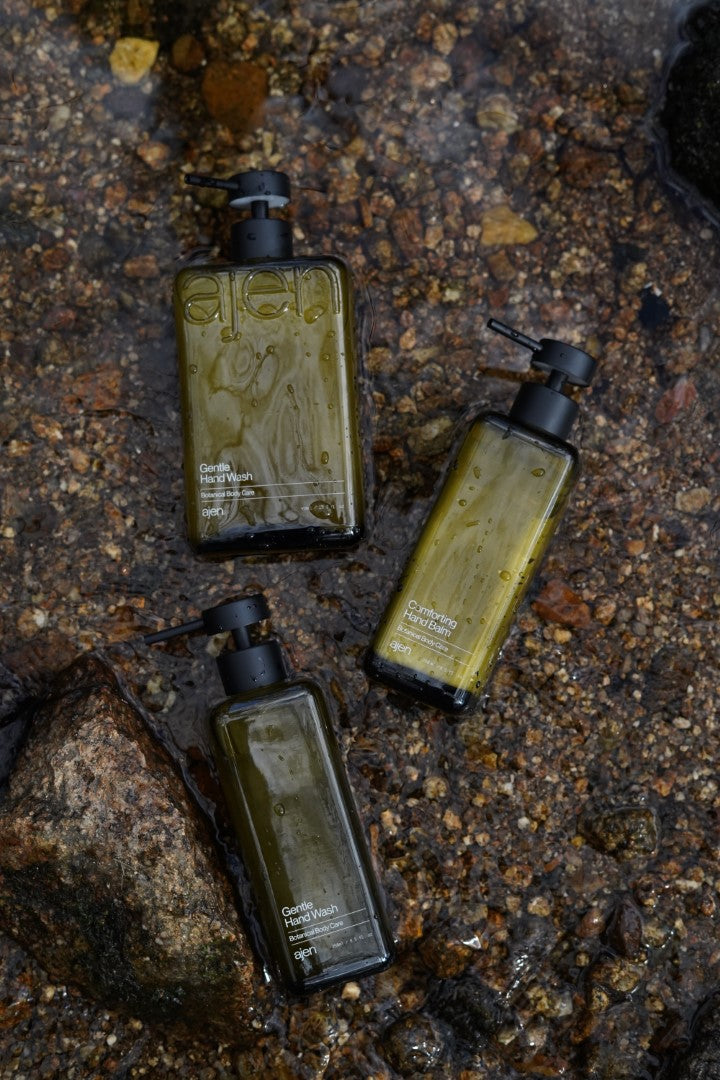Three glass Ajen bottles styled sitting in a natural water source with rocks underneath.