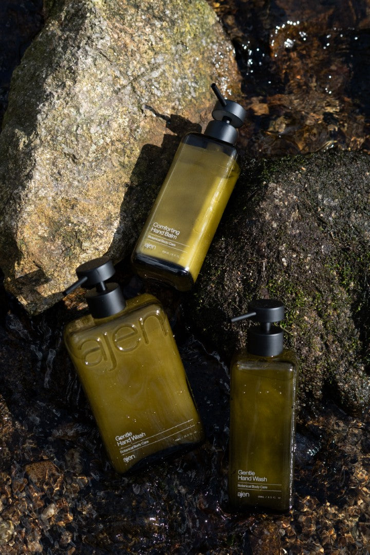 Three glass Ajen bottles styled sitting against rocks in a natural water source with rocks underneath.
