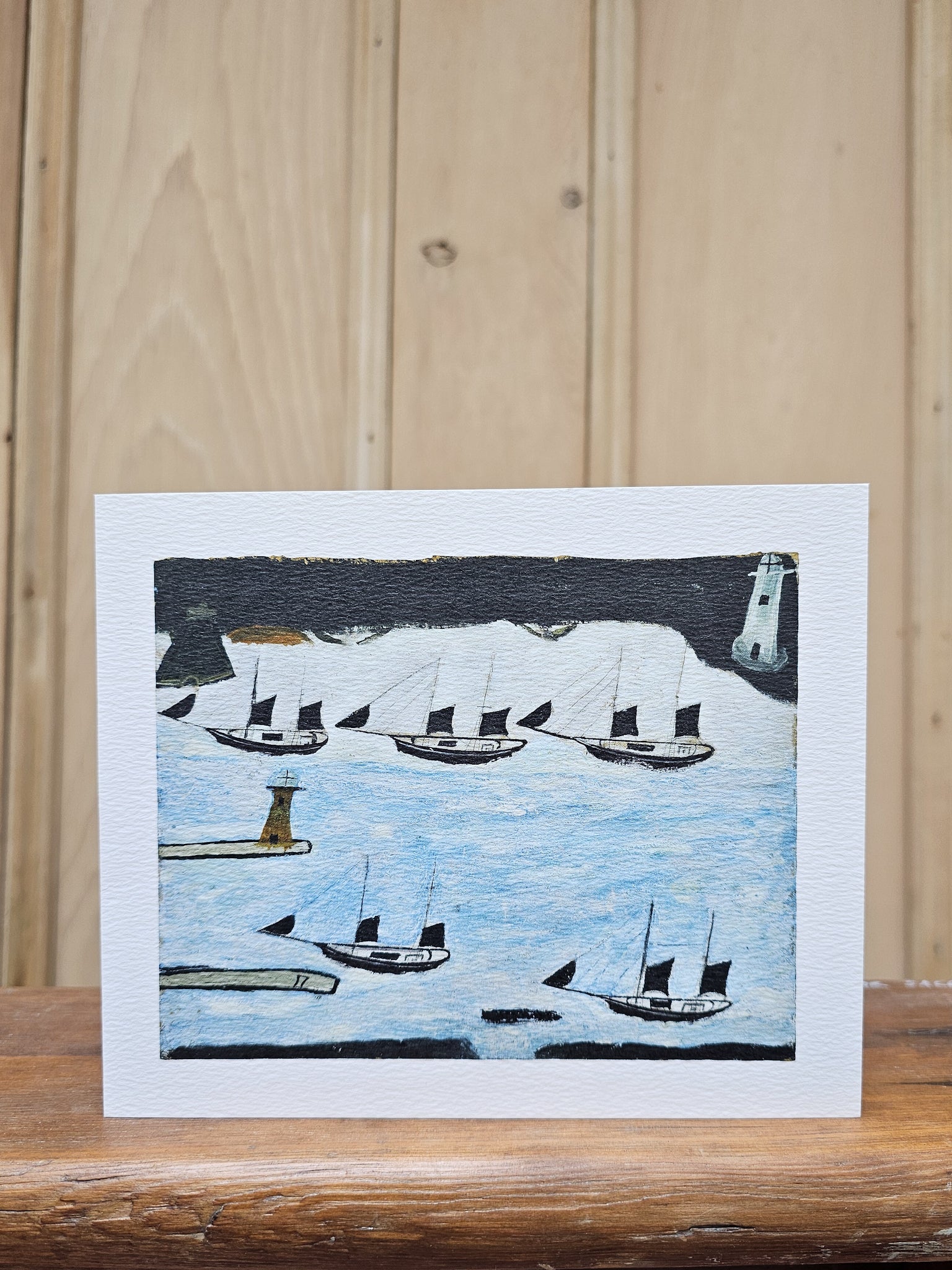Greeting card reproducing Alfred Wallis's naive painting of five sailing boats in St Michael's Mount Bay, with two lighthouses and a dark stormy sky