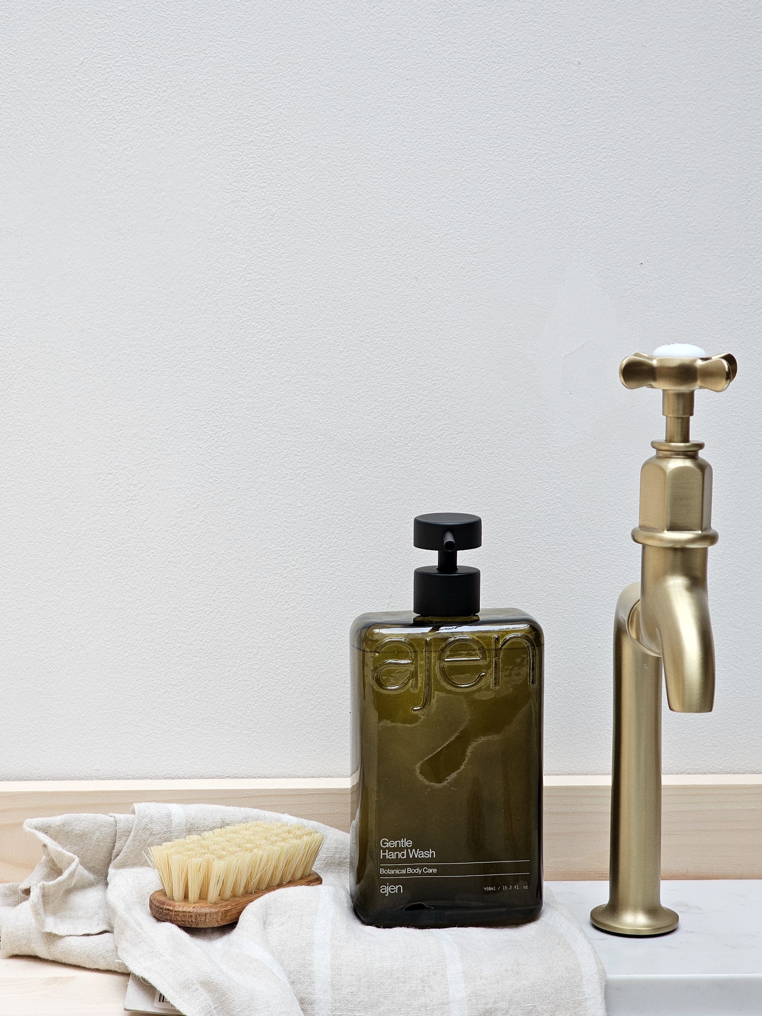 A bottle of Ajen hand wash next to a gold-colored faucet, with a white cloth and a wooden brush in the foreground.