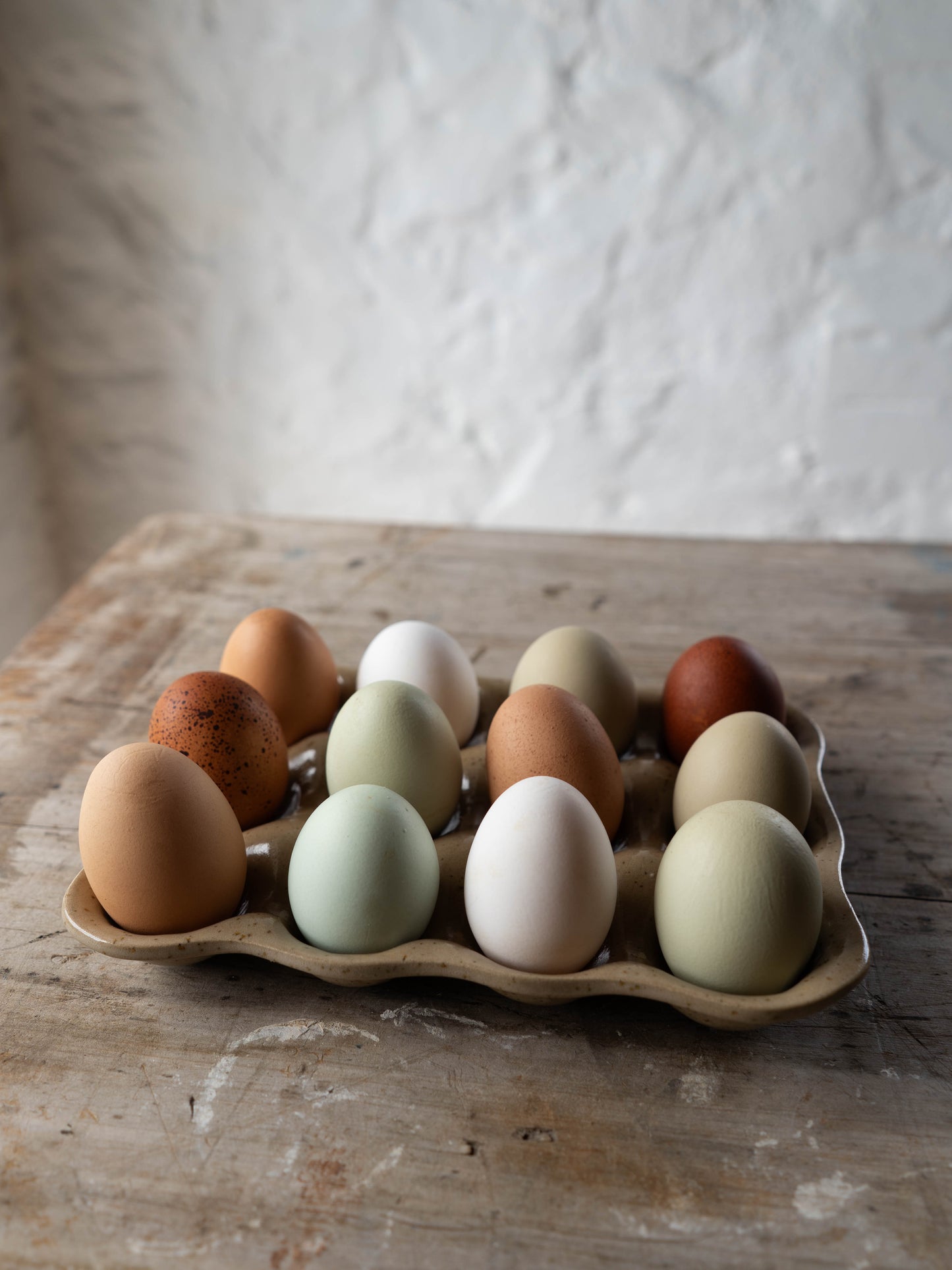 A variety of eggs with different colors and patterns placed in a stoneware egg tray on a wooden surface with a plain wall in the background.