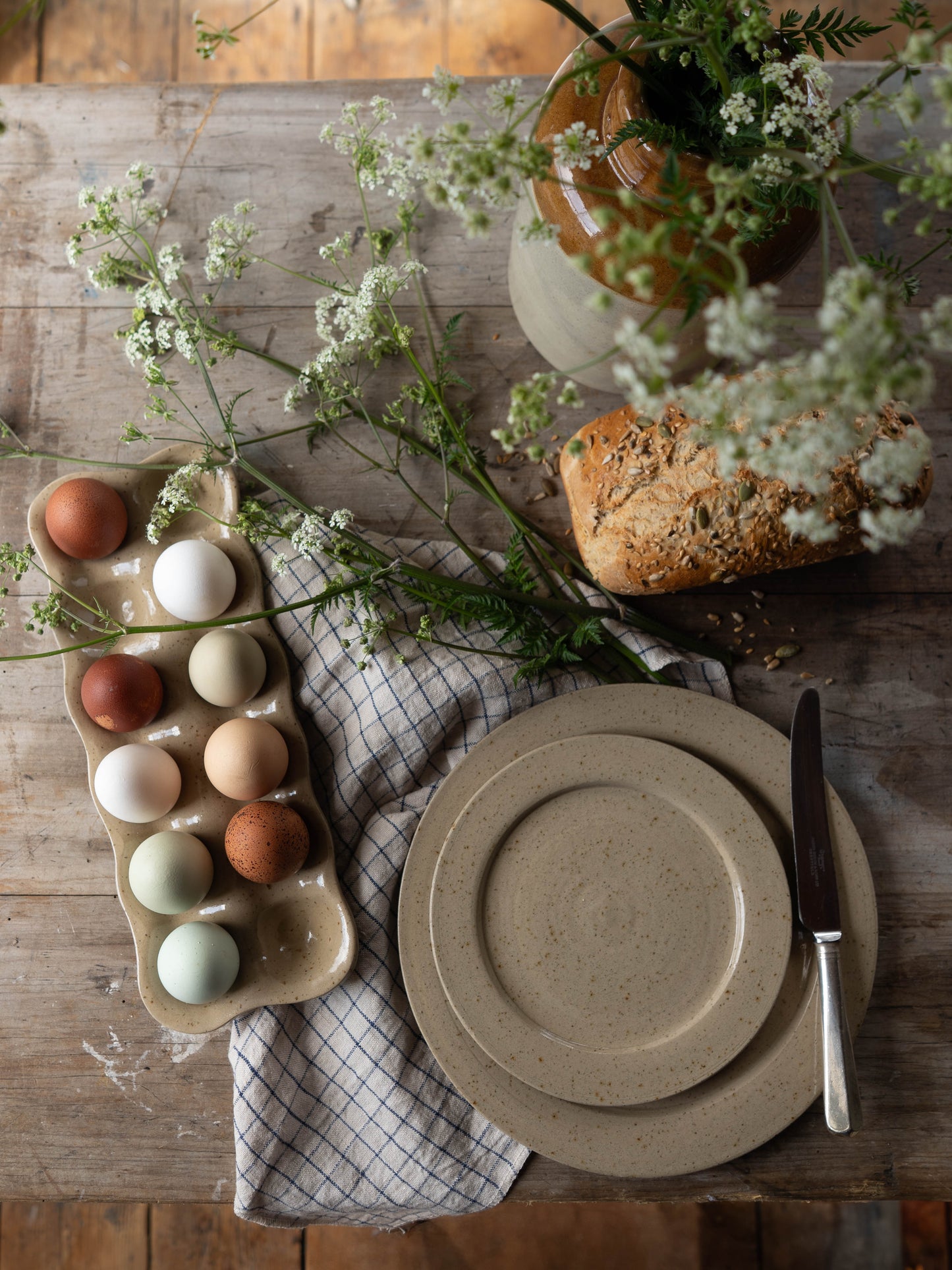 Flat lay of a rustic kitchen setting, showing a loaf of grain bread, flowers and handmade stoneware plates and handmade stoneware egg tray. 