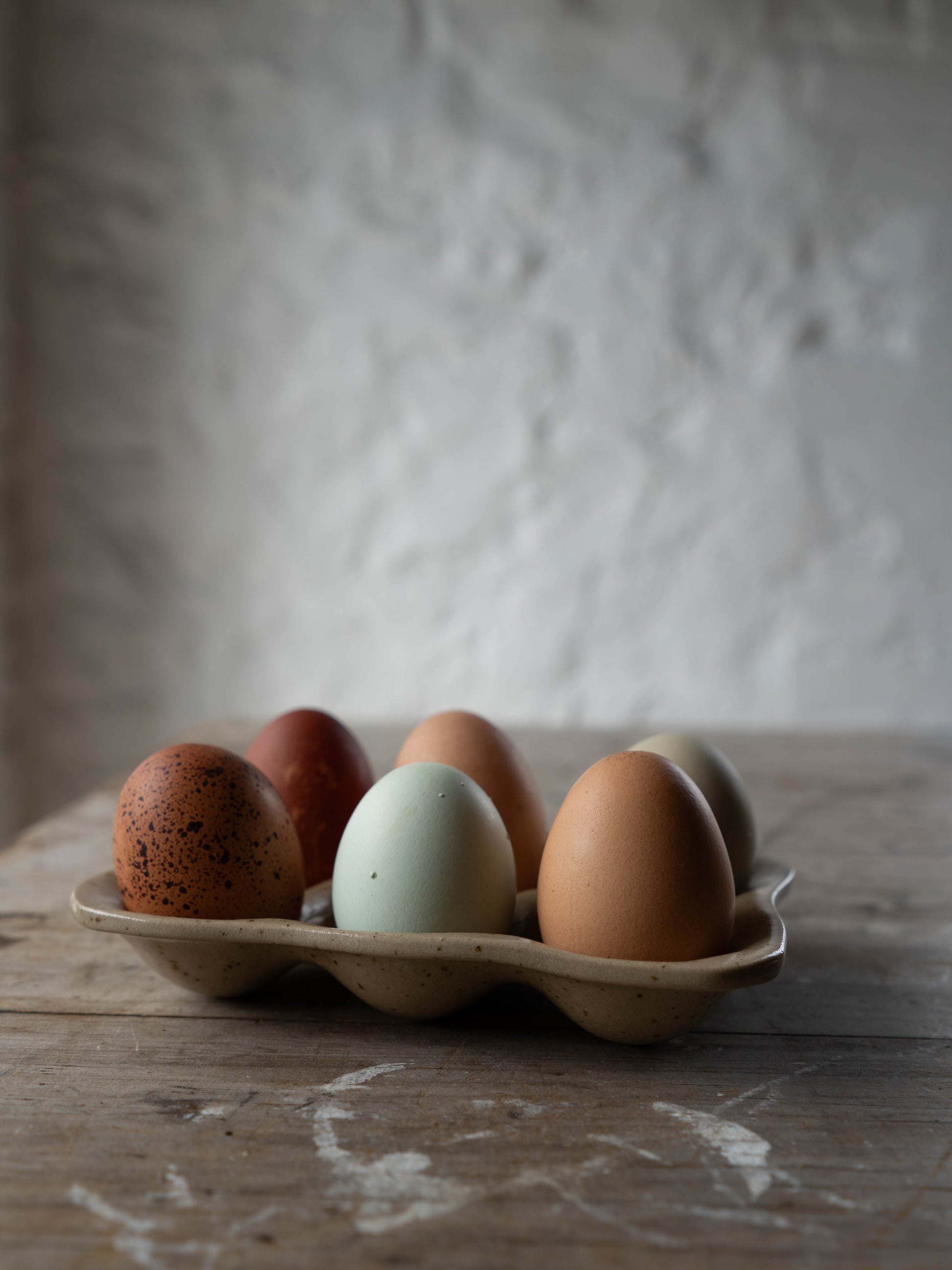 A variety of eggs with different colors and patterns placed in a stoneware egg tray on a wooden surface with a plain wall in the background.