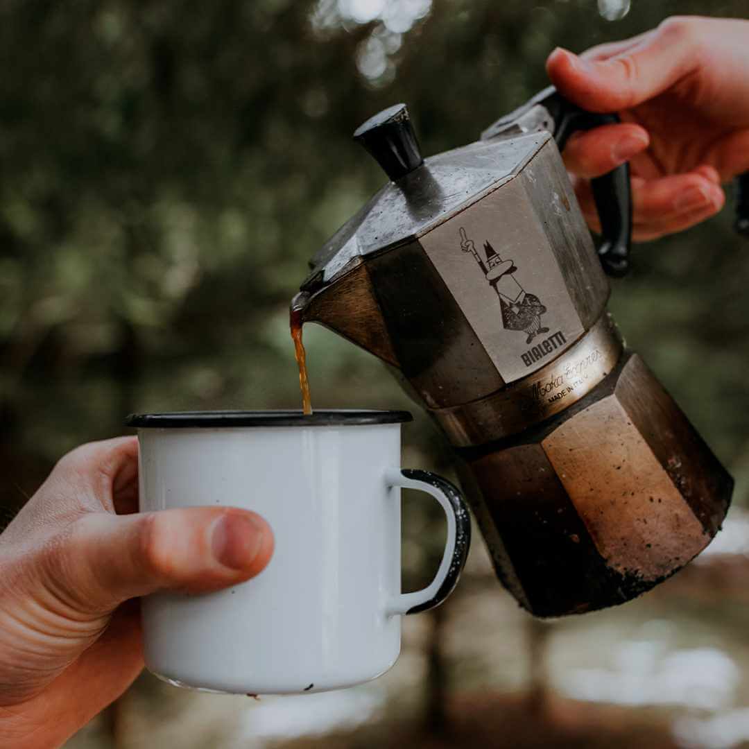 Person pouring coffee from a portable coffee maker into a white mug with a forest background.