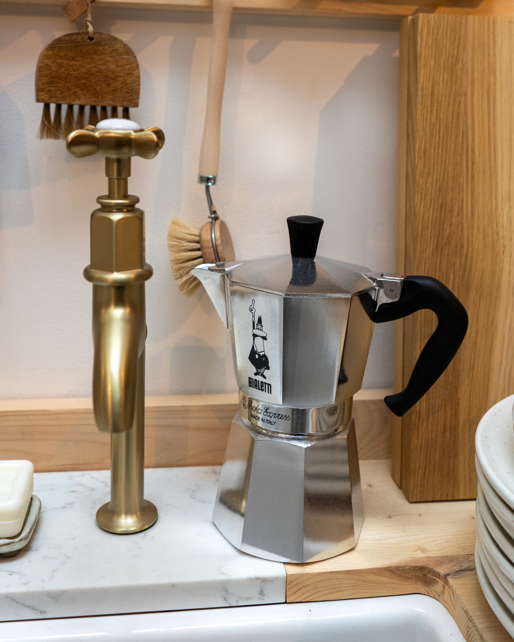 Silver Moka pot with black handle on a wood and marble worktop with cleaning brushes in the background.