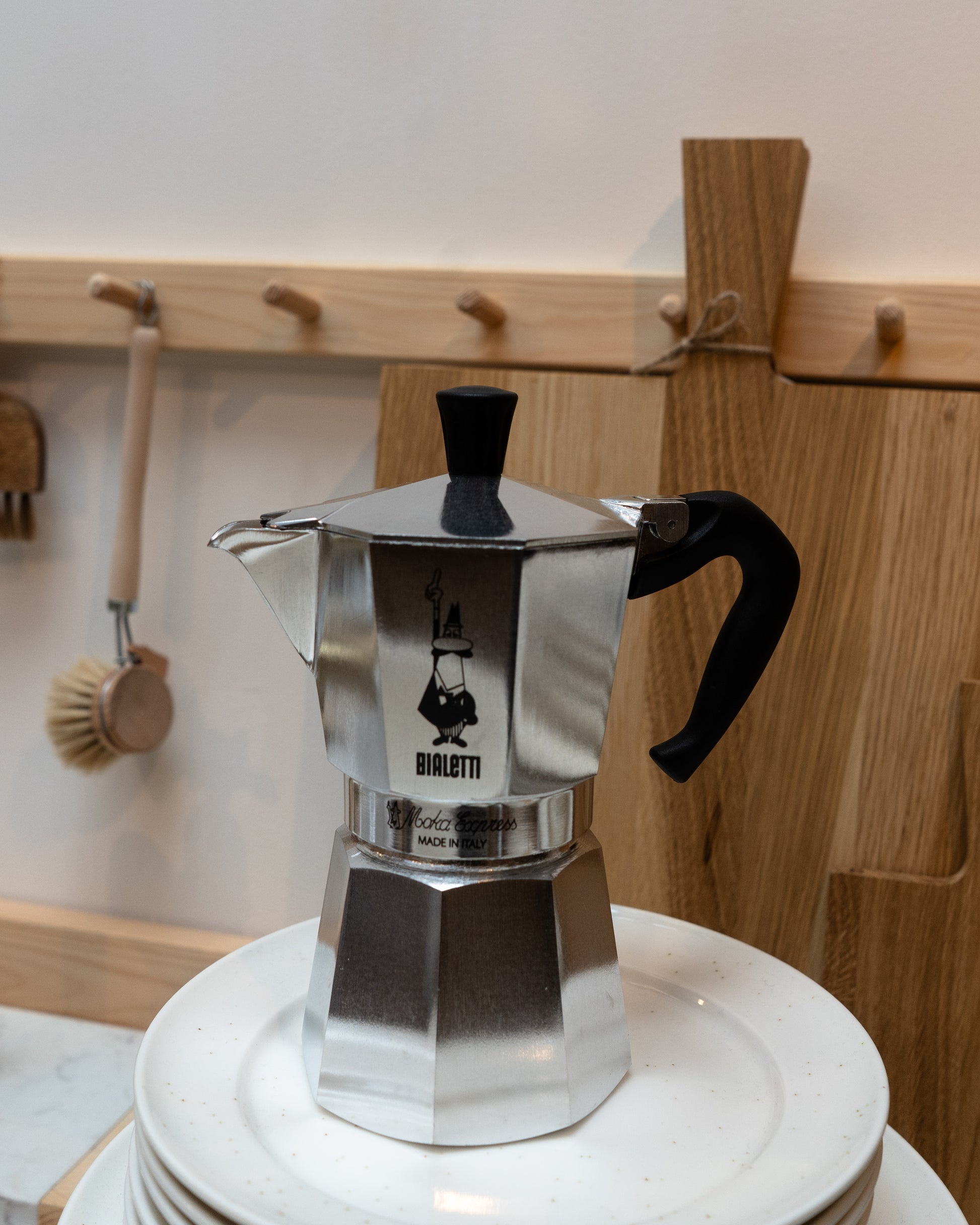 Silver Bialetti coffee maker on a white surface with wooden wall and hooks in the background