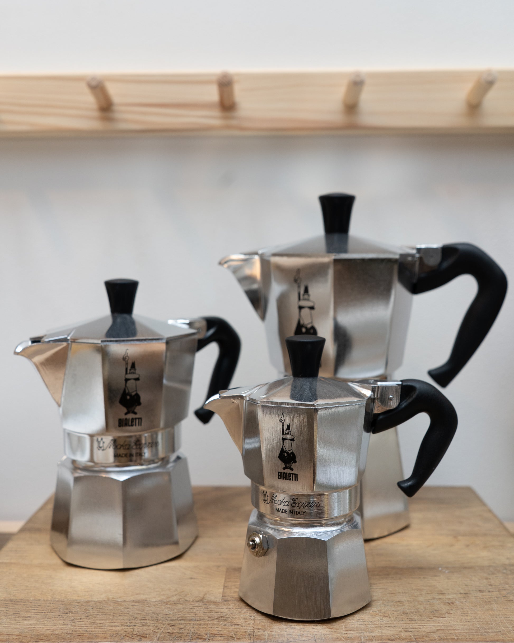 Three Bialetti Moka Express coffee makers on a wooden surface with a light-colored wall background.