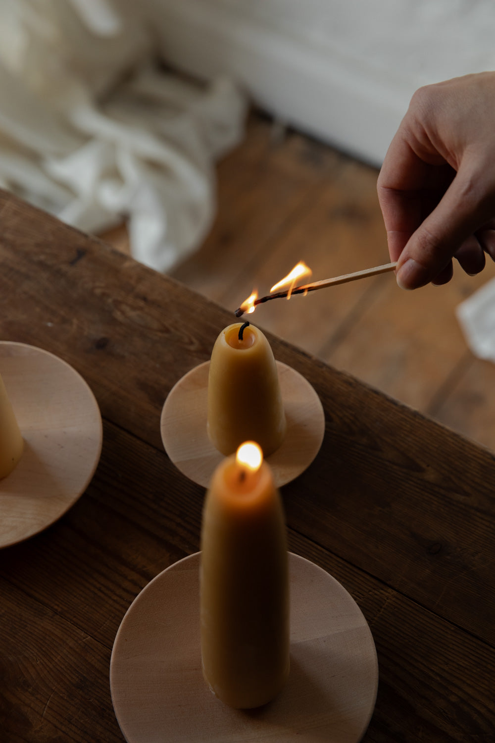 Person lighting a beeswax candle with a match sitting on a Birch wood plate on a wooden surface.