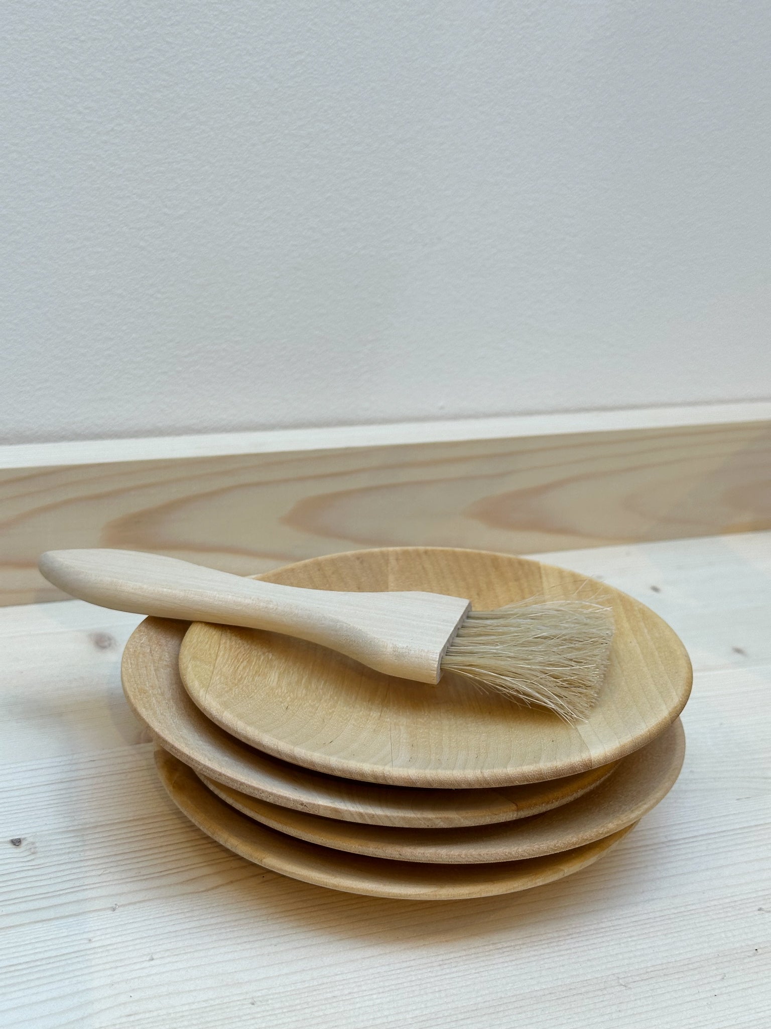 A set of four birch wood plates stacked on top of each other against a white background, with a baking brush on top.