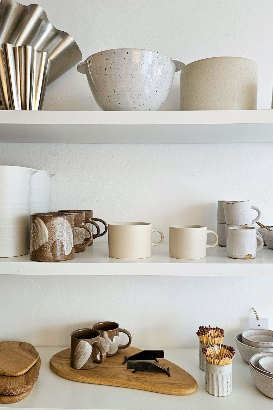 White open shelves displaying a collection of ceramic mugs, bowls, plates, and serving dishes in neutral beige, cream, and brown tones.