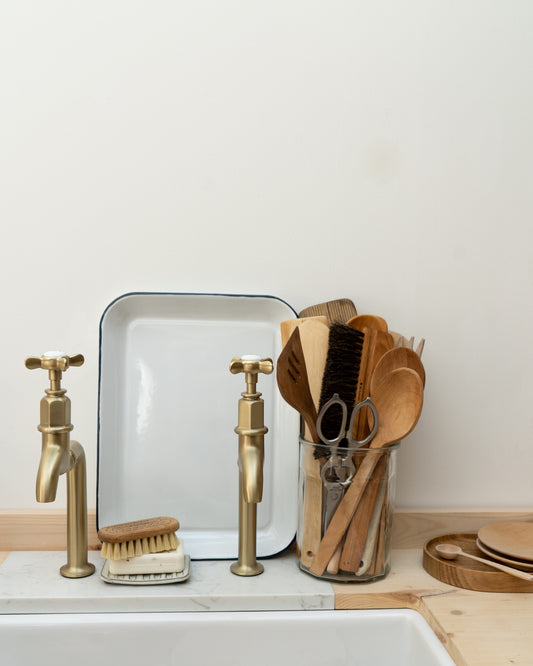 Kitchen sink with brass taps, enamel tray, scrubber, and wooden utensils on a light background