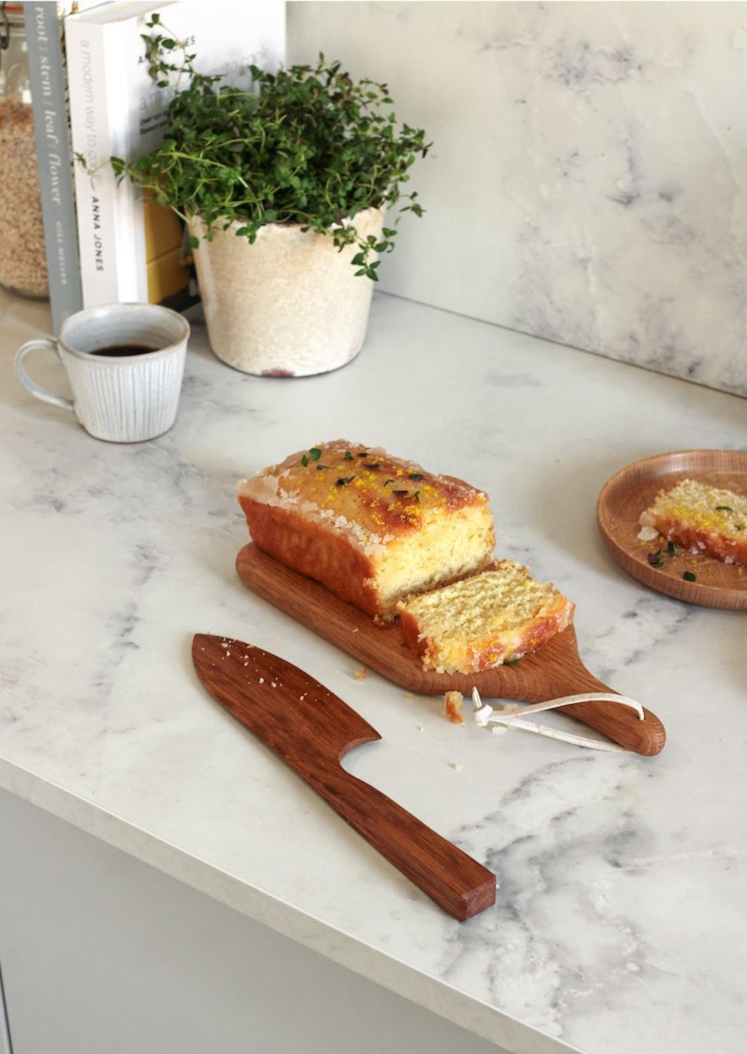 A brown English Oak cake knife is placed on a marble kitchen top. Next to the knife is a small a mini English Oak serving board with a loaf of cake with a slice cut. Nearby sits a potted plant and a hot drink in a mug.