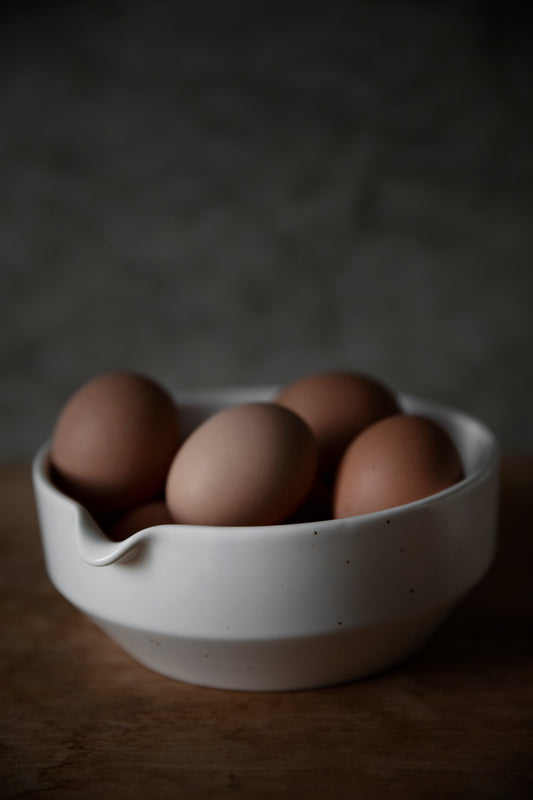 A white stoneware pouring bowl with a spout, filled with brown eggs, placed on a wooden surface.