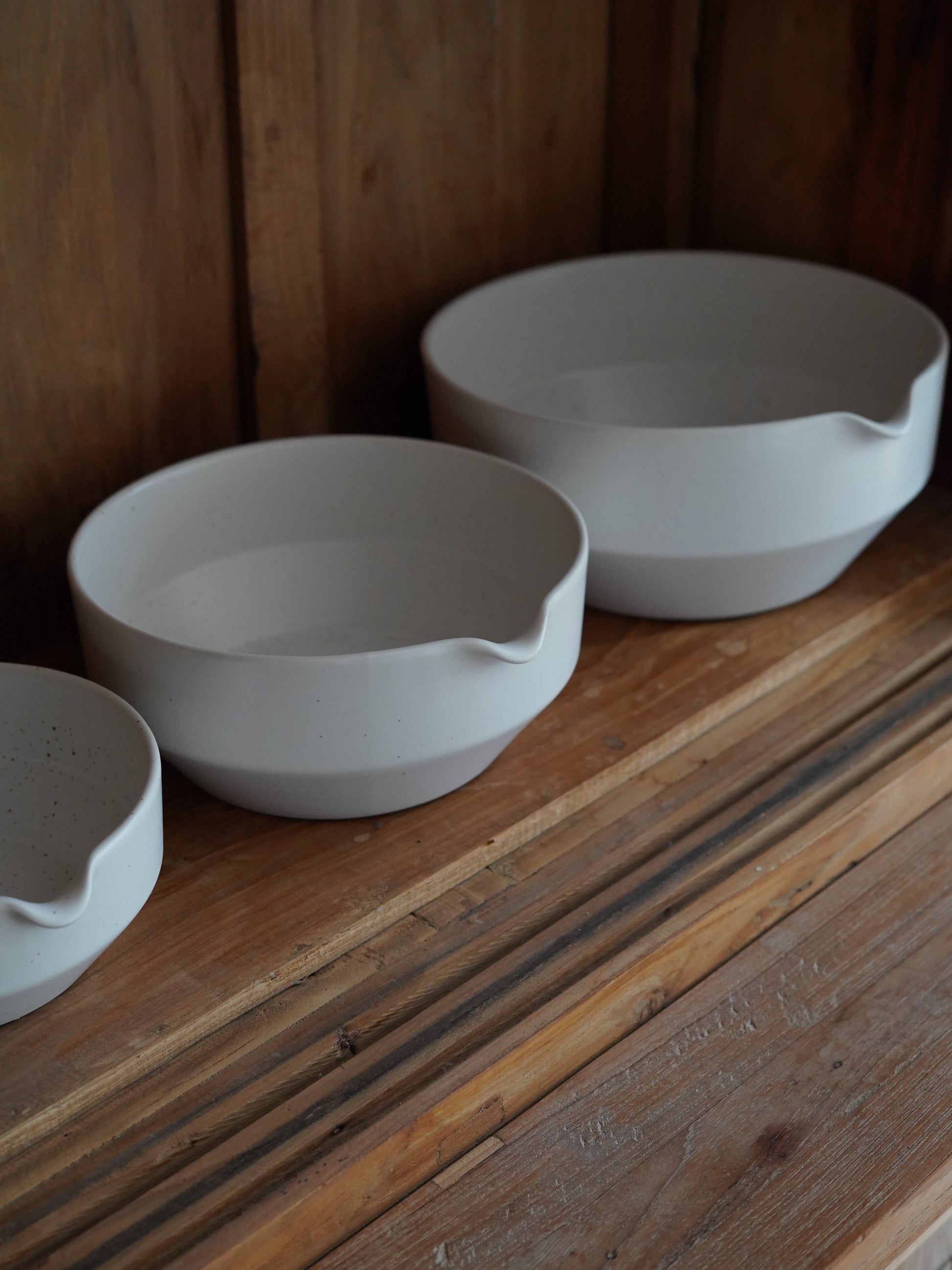 Three minimalist white ceramic pouring bowls in graduated sizes are arranged on a rustic wooden shelf. Each bowl features a soft, organic shape with a subtle spout, blending modern simplicity with functional design. The warm wood backdrop enhances the understated elegance of the set.