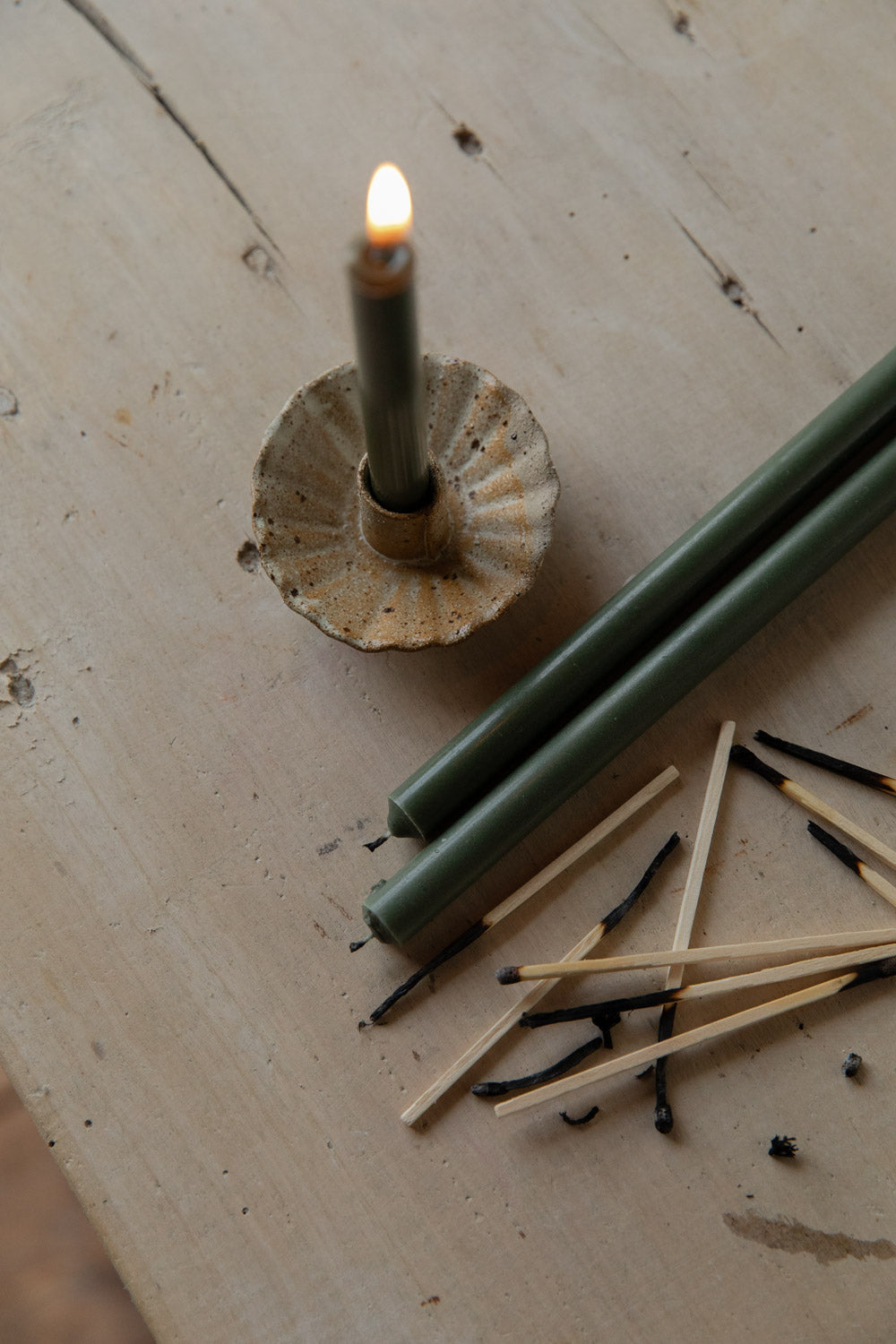 Green candle in a fluted natural candle holder with a lit wick on a wooden surface with matches nearby.