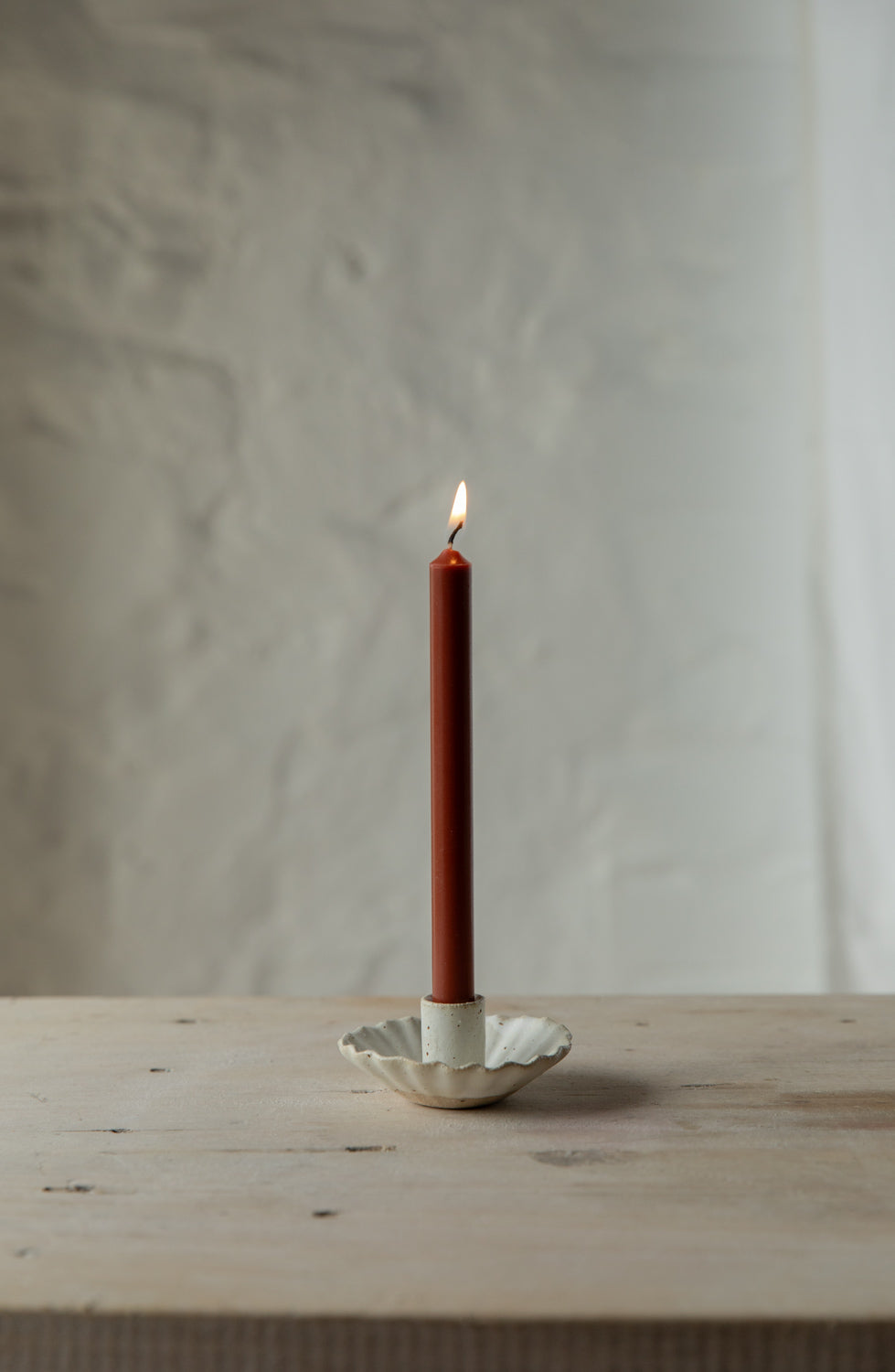 Red candle in a handmade fluted white candle holder on a wooden surface with a neutral background.