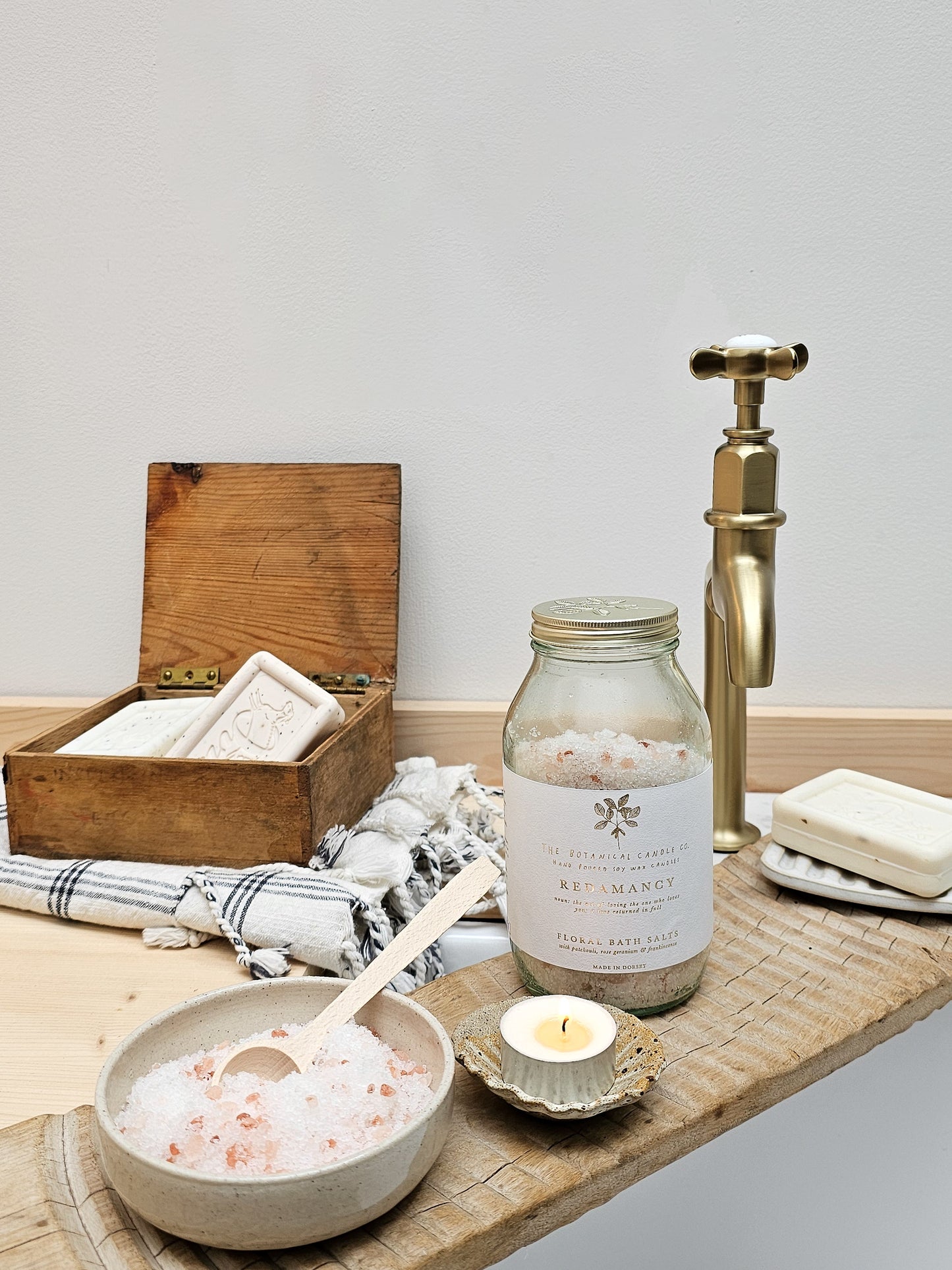 Bath salts in a jar, bowl, and wooden box on a wooden surface with a candle and soap.