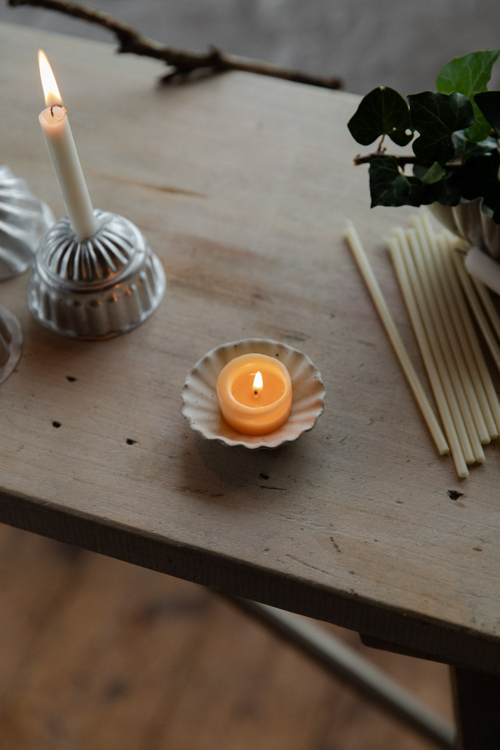 Candle in a fluted white dish on a wooden surface with other candles and a plant in the background.