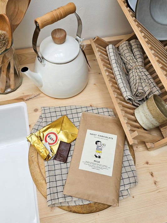 White teapot with wooden handle on a wooden surface with a package of Heist 
Chocolate and a bar of chocolate.