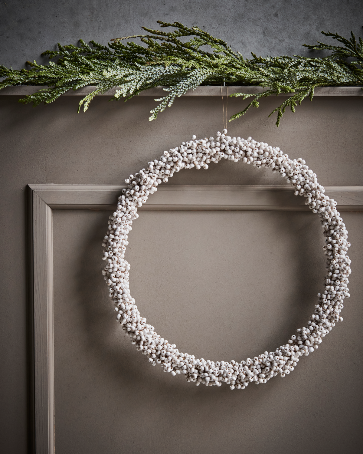 White wooden bead Christmas wreath hanging against a panelled wall, with a branch of fir tree resting on a shelf above.