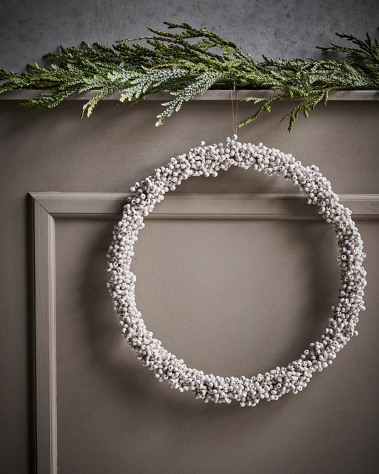 White wooden bead Christmas wreath hanging against a panelled wall, with a branch of fir tree resting on a shelf above.
