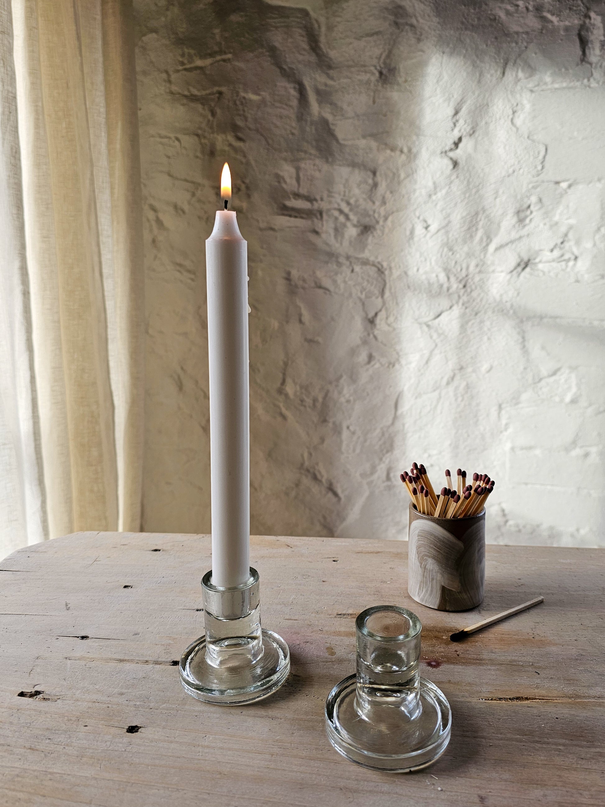 Two glass candle holders, with white candles on a wooden table with a textured white background.