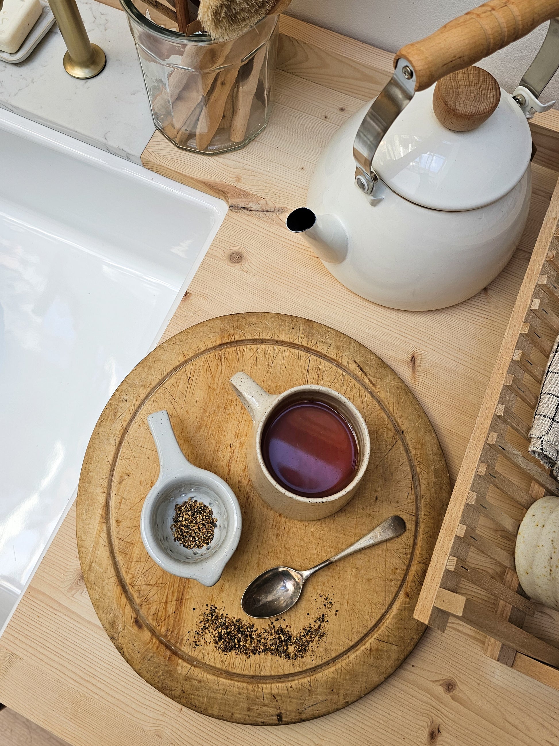 Tea-making setup with a teapot, ceramic tea strainer, cup of tea, and wooden board on a kitchen counter.