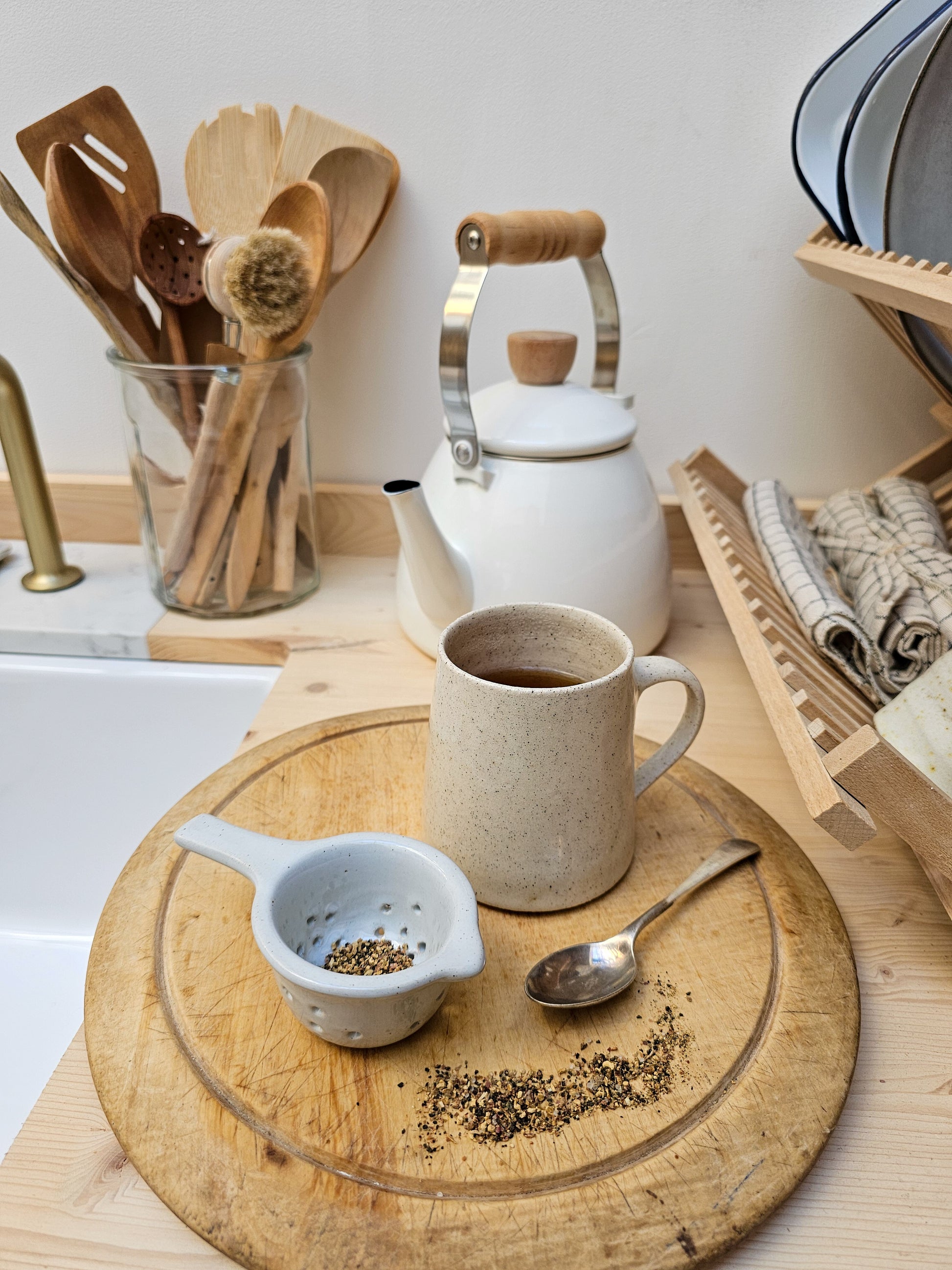 Tea-making setup with a teapot, ceramic tea strainer, cup of tea, and wooden board on a kitchen counter.