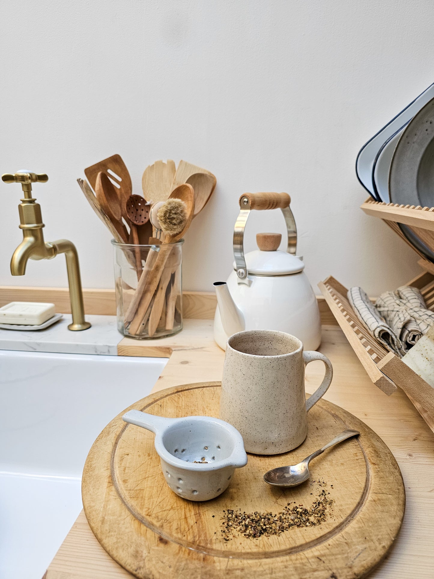 Tea-making setup with a teapot, ceramic tea strainer, cup of tea, and wooden board on a kitchen counter.
