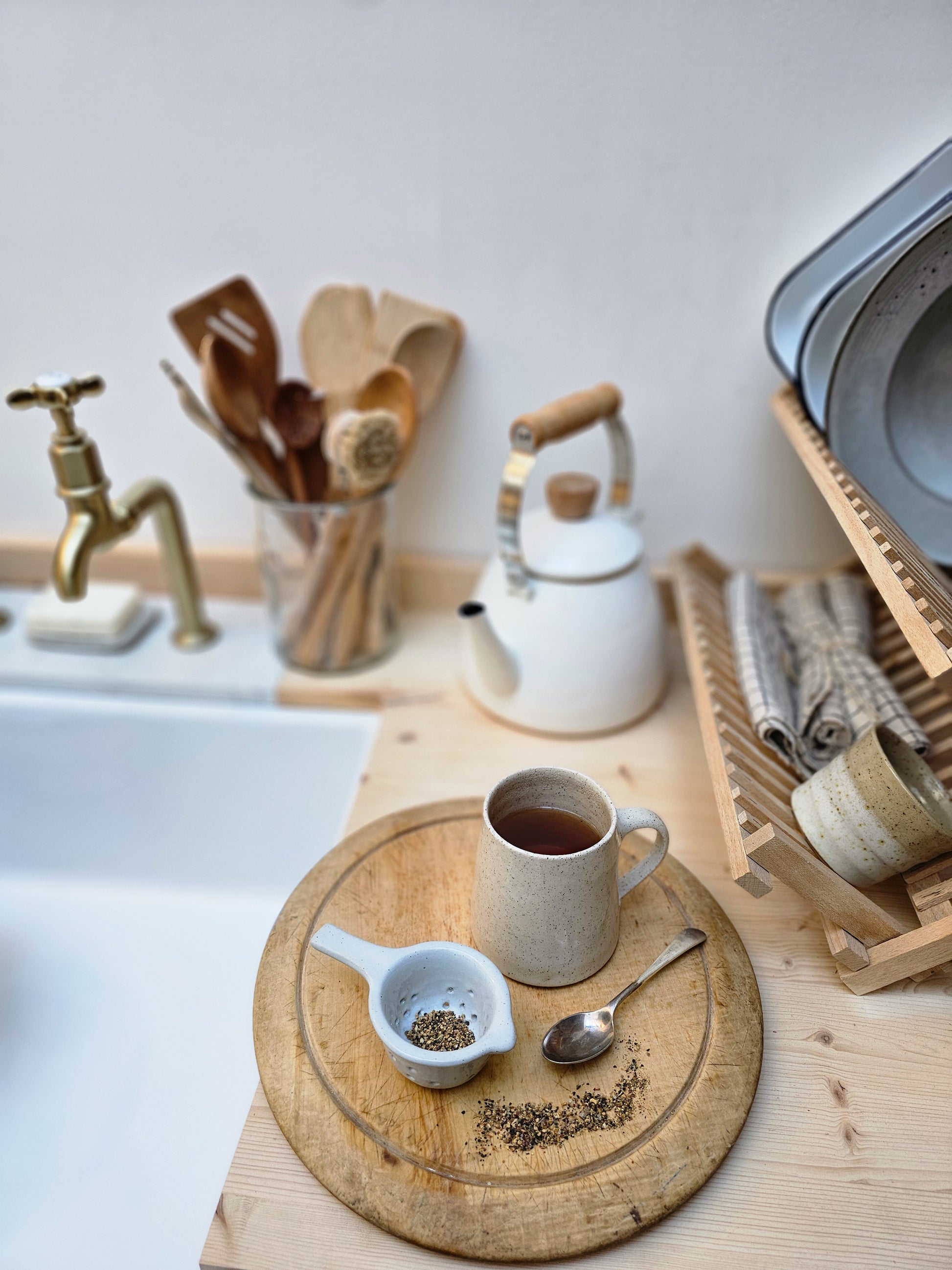 Tea-making setup with a teapot, ceramic tea strainer, cup of tea, and wooden board on a kitchen counter.