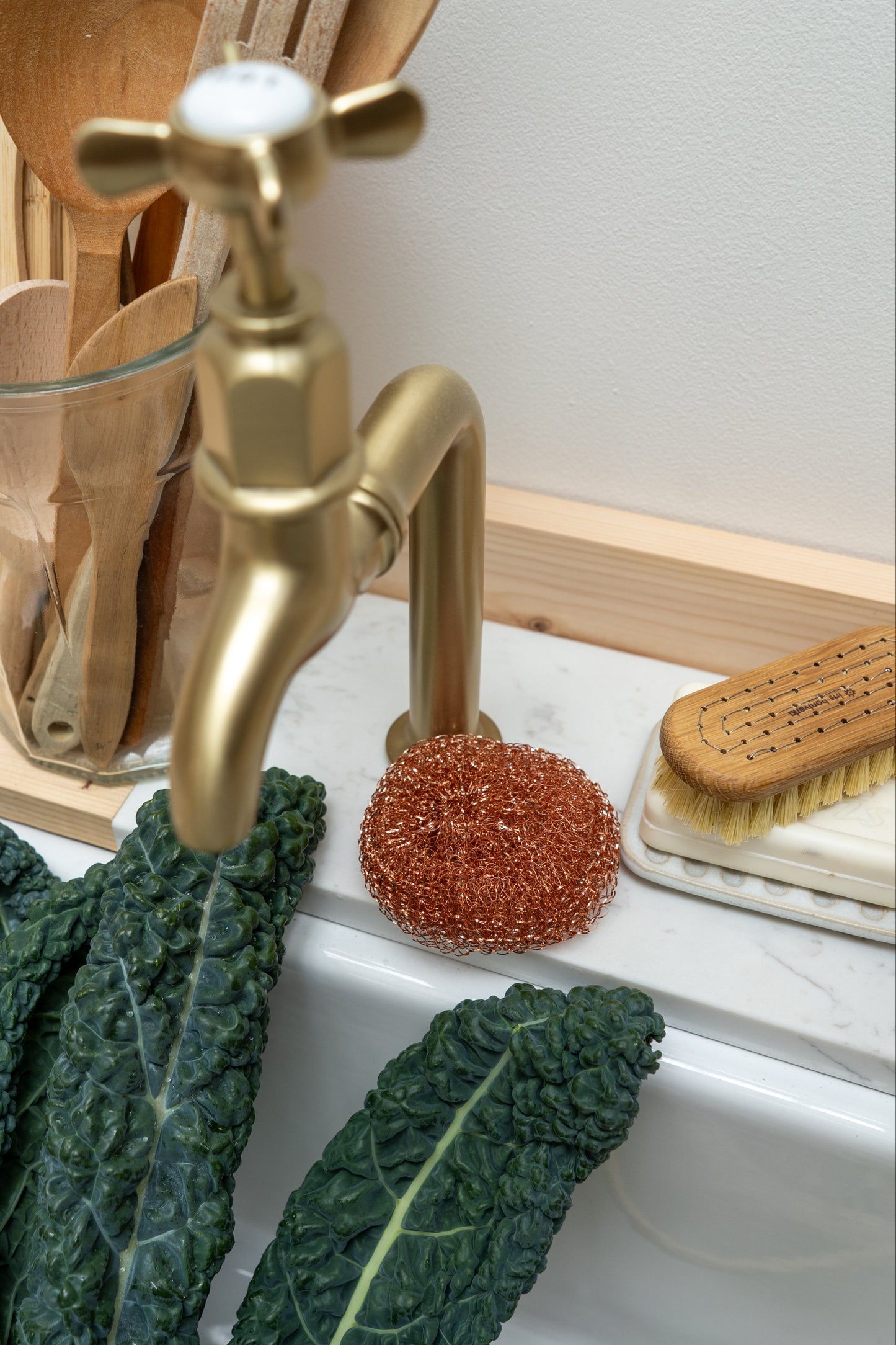 Gold faucet with kitchen utensils and vegetables on a white surface, next to a wooden nail brush.