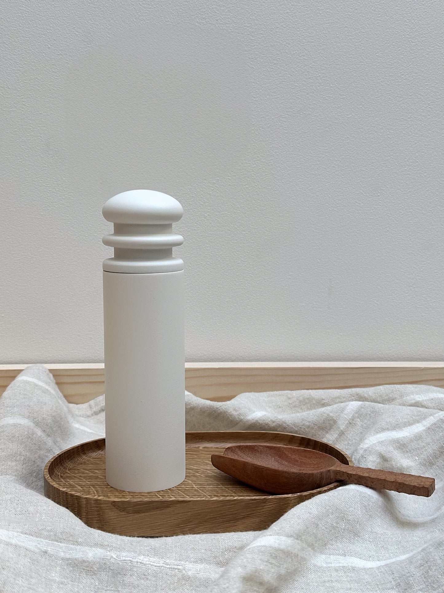A white ceramic salt/pepper mill on a wooden tray with a wooden spoon beside it, against a neutral background.