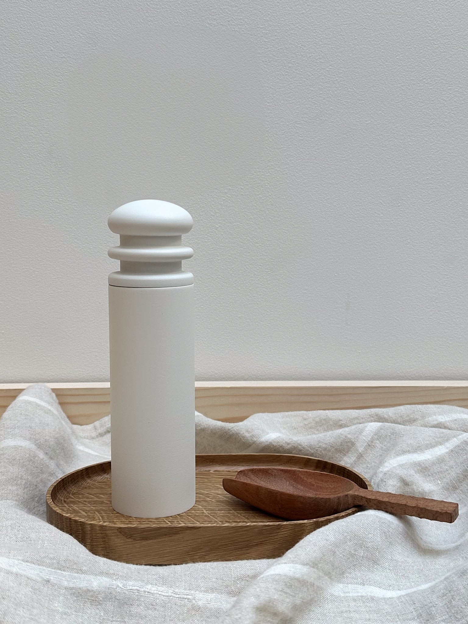 A white ceramic salt/pepper mill on a wooden tray with a wooden spoon beside it, against a neutral background.