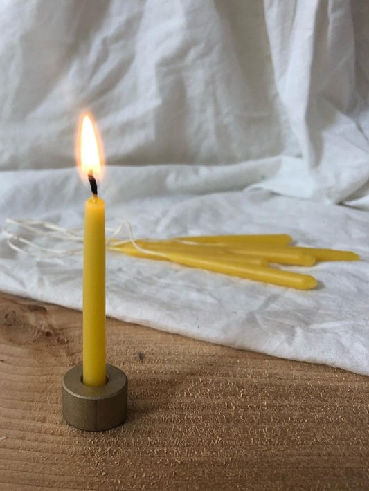 A yellow beeswax mini candle lit, placed on a grey holder with several unlit candles behind it, set on a wooden surface with a white cloth in the background.
