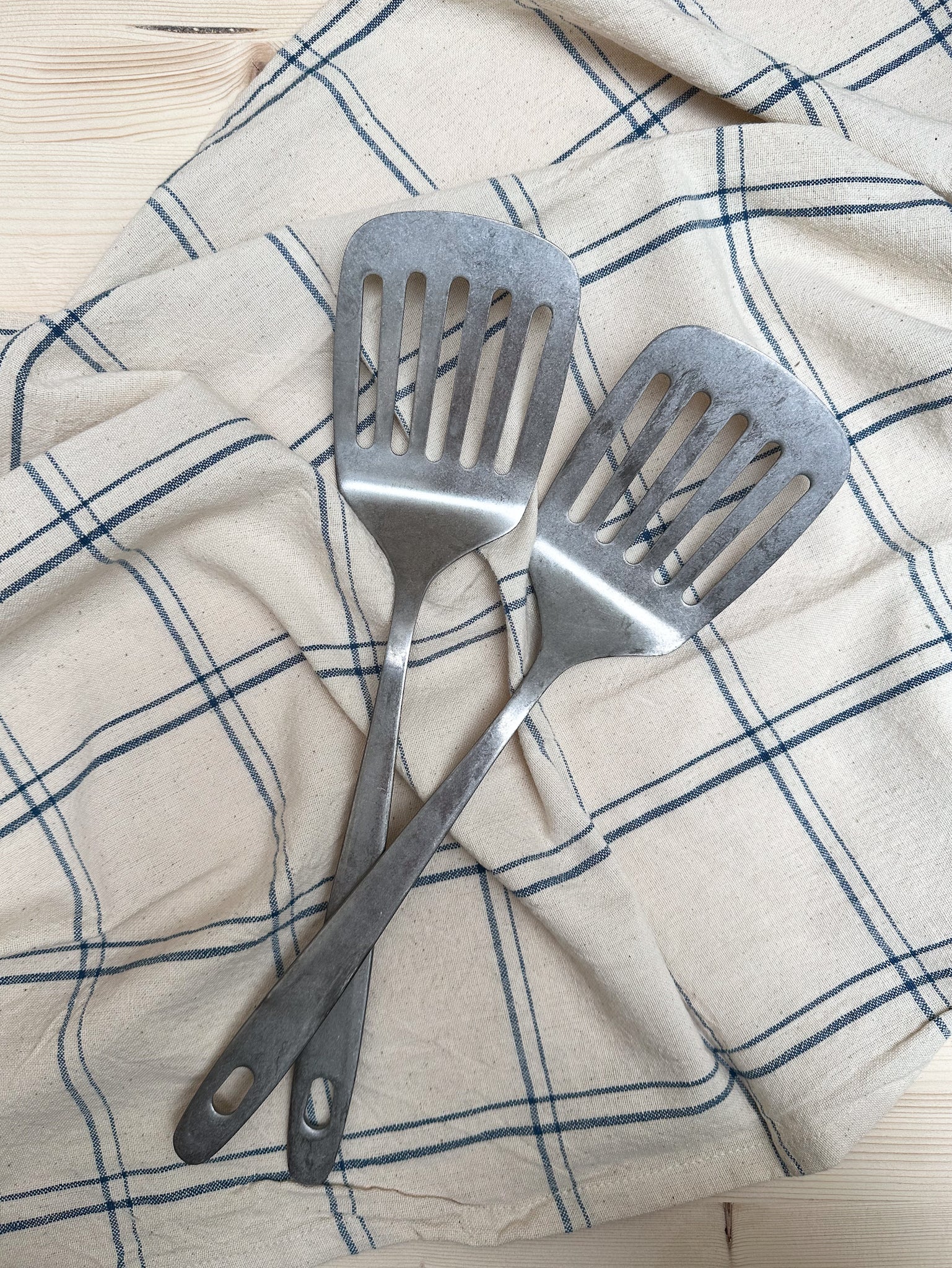 Two stainless steel slotted spatulas placed on a kitchen towel with a checkered pattern