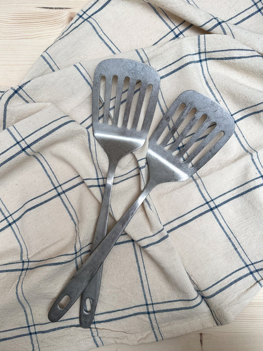 Two stainless steel slotted spatulas placed on a kitchen towel with a checkered pattern