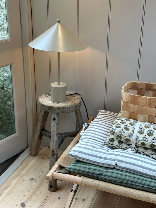 A minimal, cone-shaped table lamp with a matte finish sits on a rustic wooden stool beside a glass-panelled door. Nearby, a woven basket and neatly folded striped and block-printed cushions rest on a natural wood daybed with a canvas sling. The setting features pale wood flooring and vertical wall panelling, evoking a calm, Scandinavian-inspired interior.