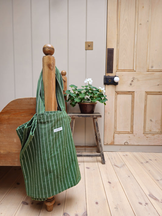 Green striped recycled cotton tote bag from OYOY hanging on a wooden bedpost, with a potted white geranium on a rustic stool and natural wood tones in the background.