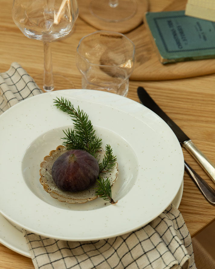 Dinner setting with a smaller scalloped natural dish placed inside the white plate, with a fig and greenery.