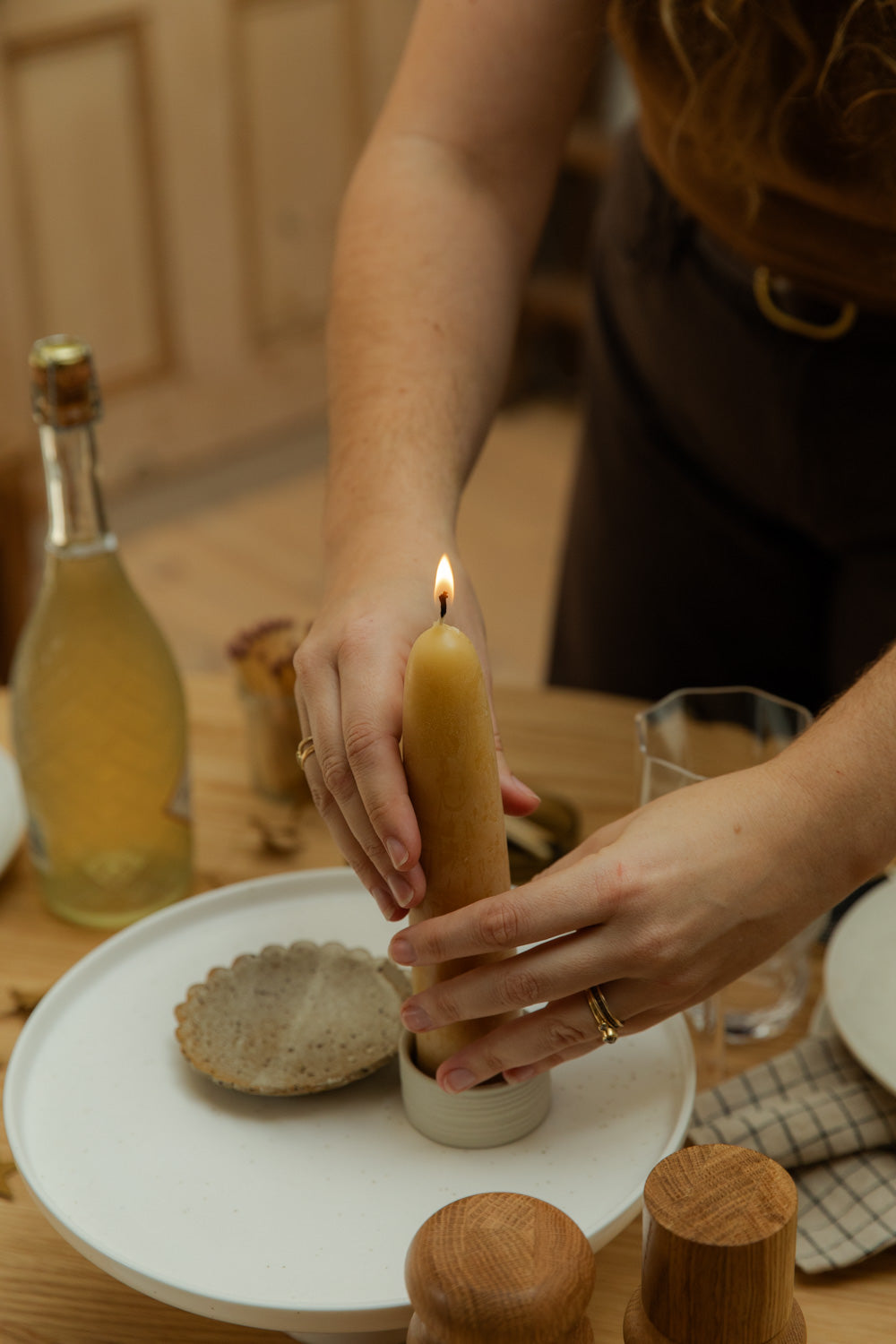 Person lighting a candle on a wooden table with a bottle and glasses in the background.
