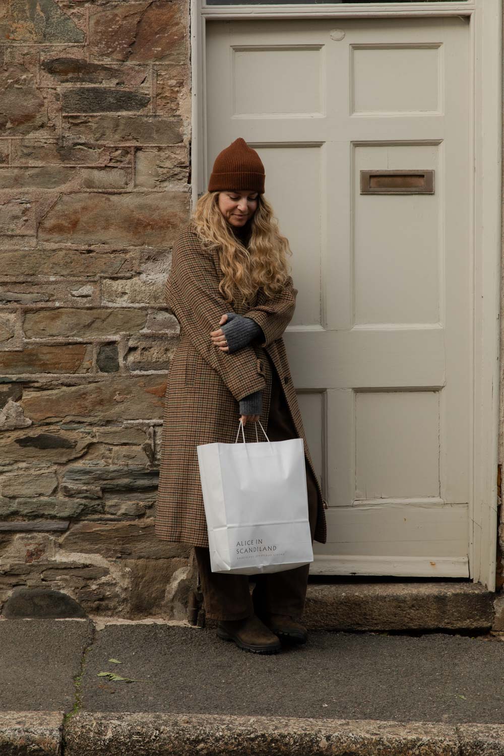 Woman in a brown ribbed beanie leaning by a painted door holding an Alice in Scandiland paper carrier bag