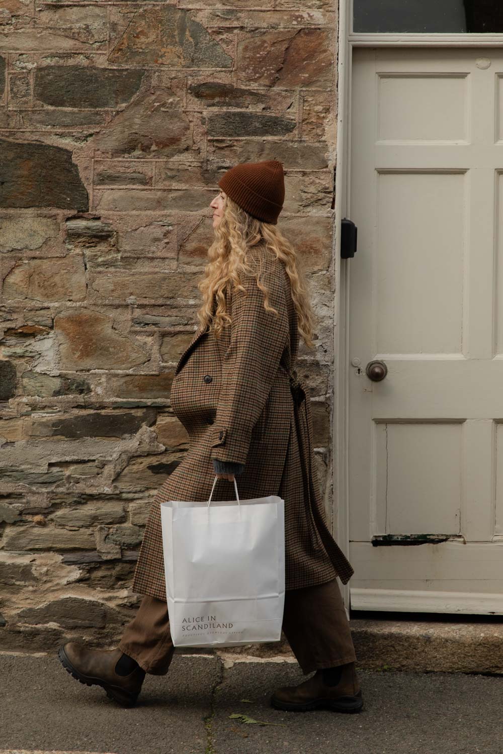 Woman in a check coat and brown ribbed beanie walking past a stone cottage with an Alice in Scandiland bag