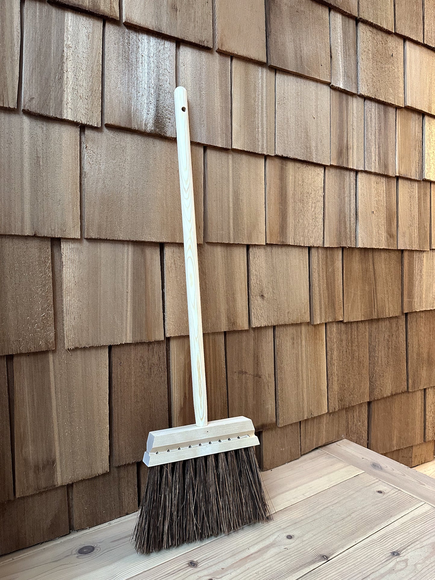 A traditional stiff bristle broom with a short wooden handle leaning against a wooden slatted wall.