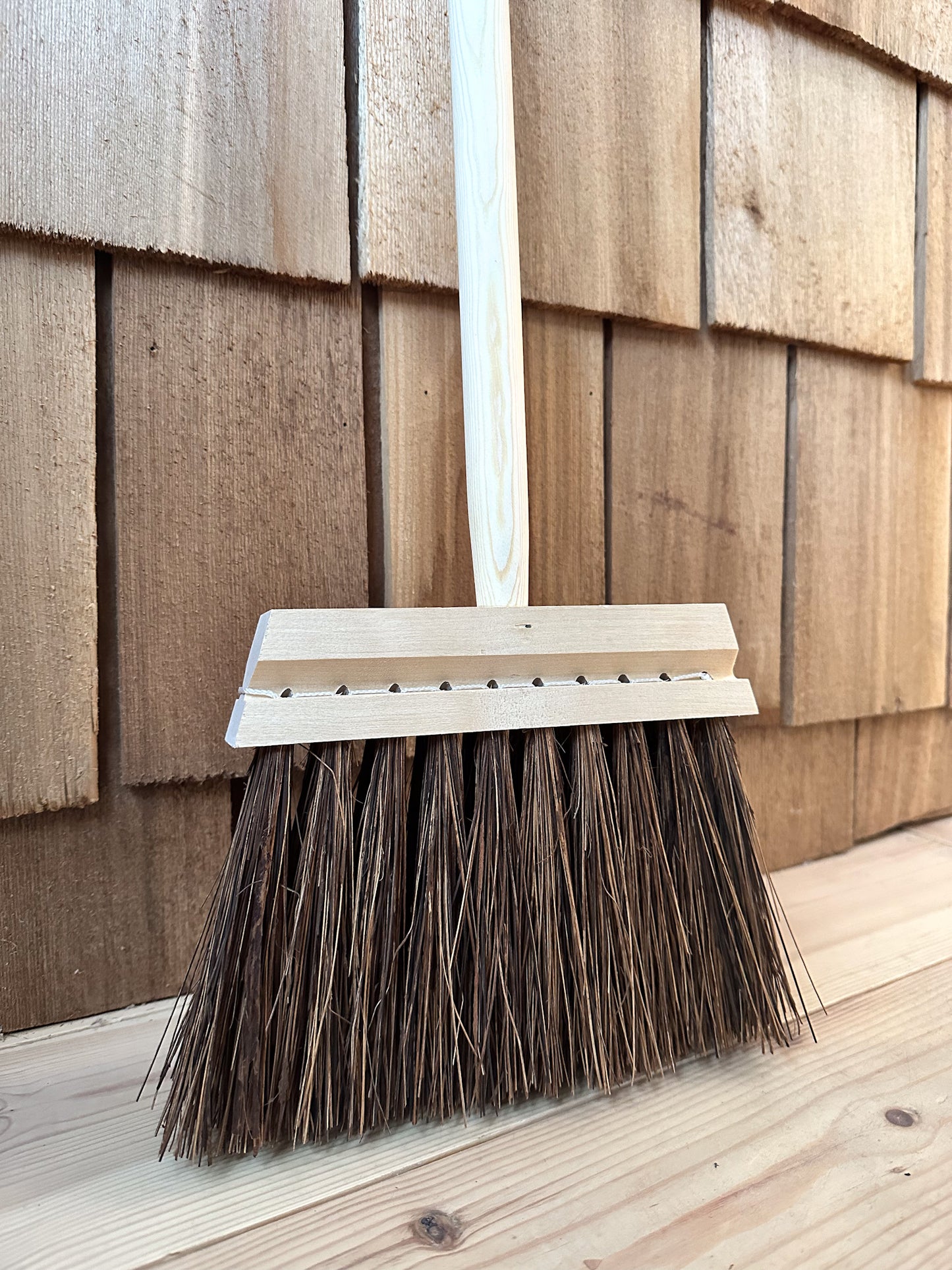 A close up of a traditional stiff bristle broom with a short wooden handle leaning against a wooden slatted wall.