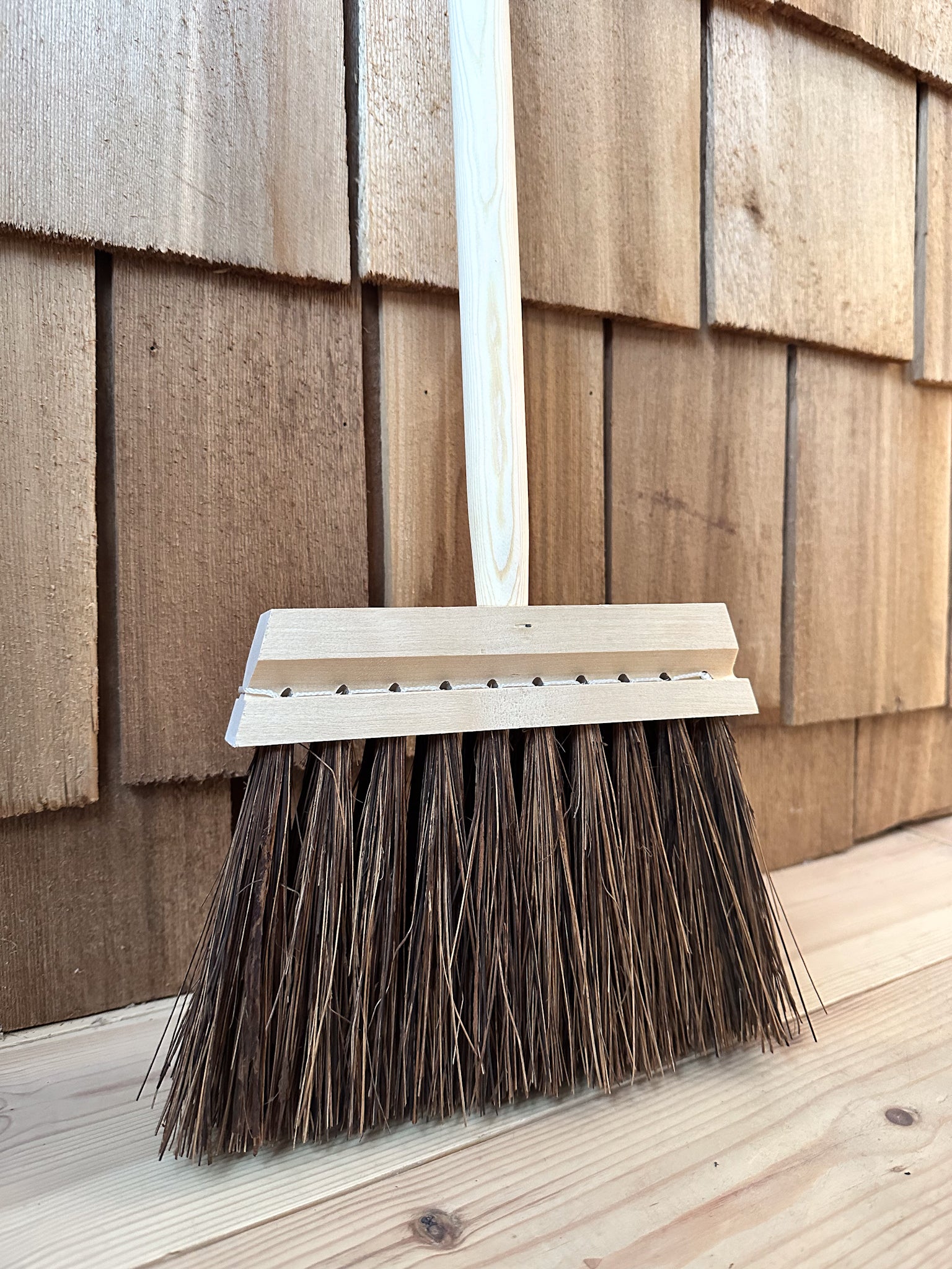 A close up of a traditional stiff bristle broom with a short wooden handle leaning against a wooden slatted wall.