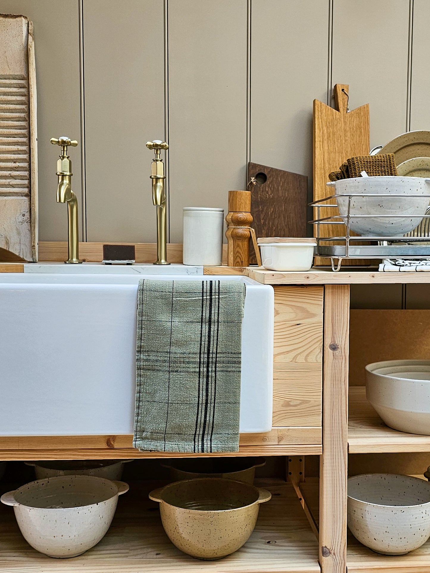 A rustic kitchen scene featuring a white farmhouse sink with brass faucets, a green plaid towel draped over the front, wooden shelves, and ceramic bowls. The background consists of beige vertical wood paneling.