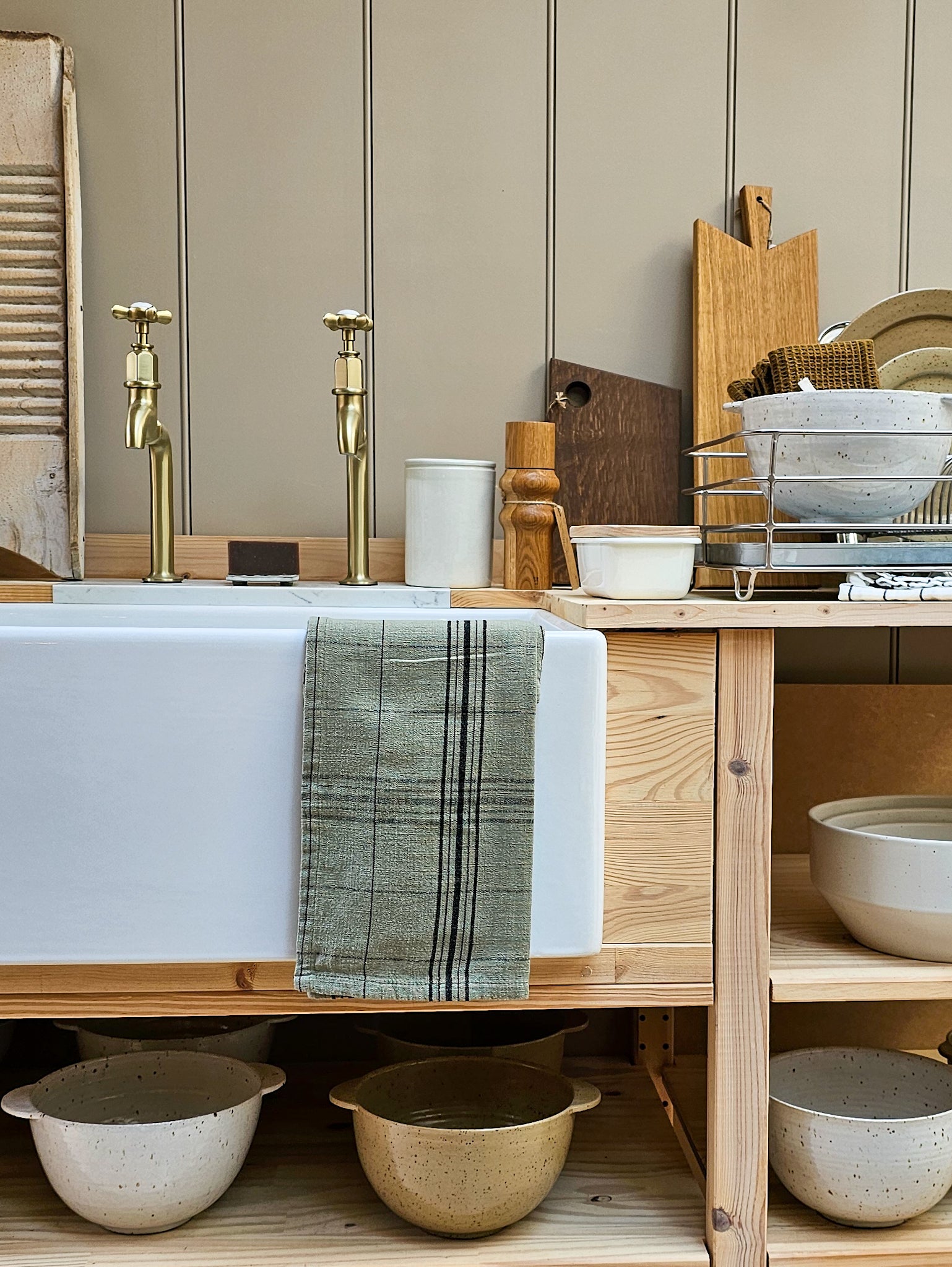 A rustic kitchen scene featuring a white farmhouse sink with brass faucets, a green plaid towel draped over the front, wooden shelves, and ceramic bowls. The background consists of beige vertical wood paneling.