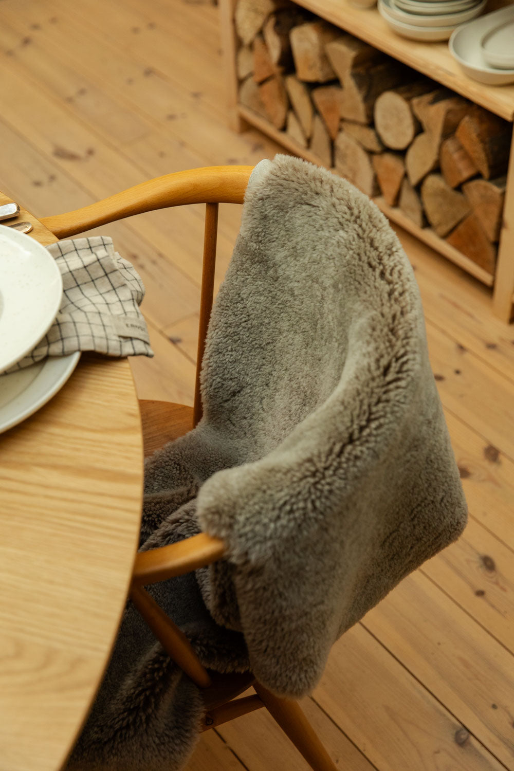 Chair with a sheepskin cover in a rustic setting with wooden floor and stacked logs.