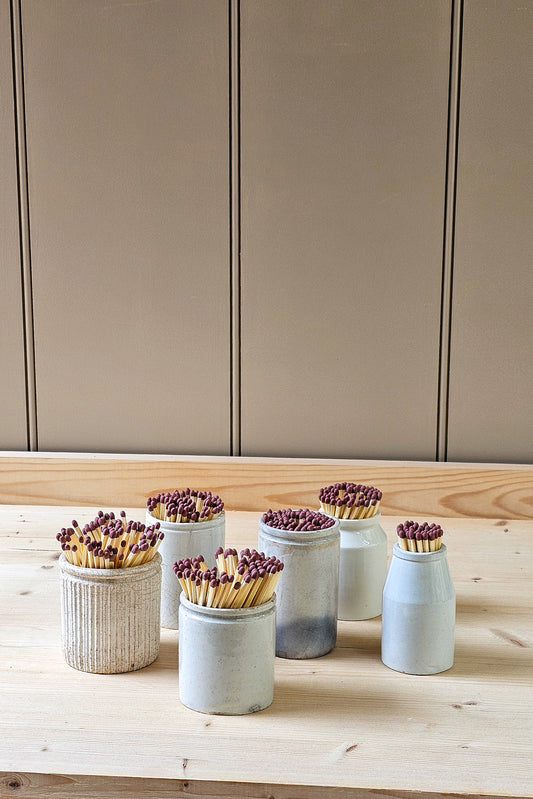Set of small vintage ceramic pots with matches on a wooden surface against a neutral wall.