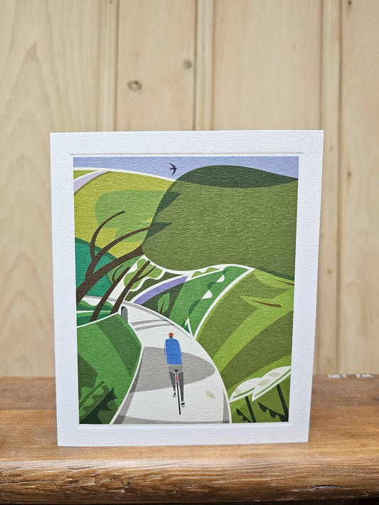 Greeting card reproducing Andrew Pavitt's illustration Lost Lanes, showing a lone cyclist on a winding country lane through lush green hills with a swift overhead